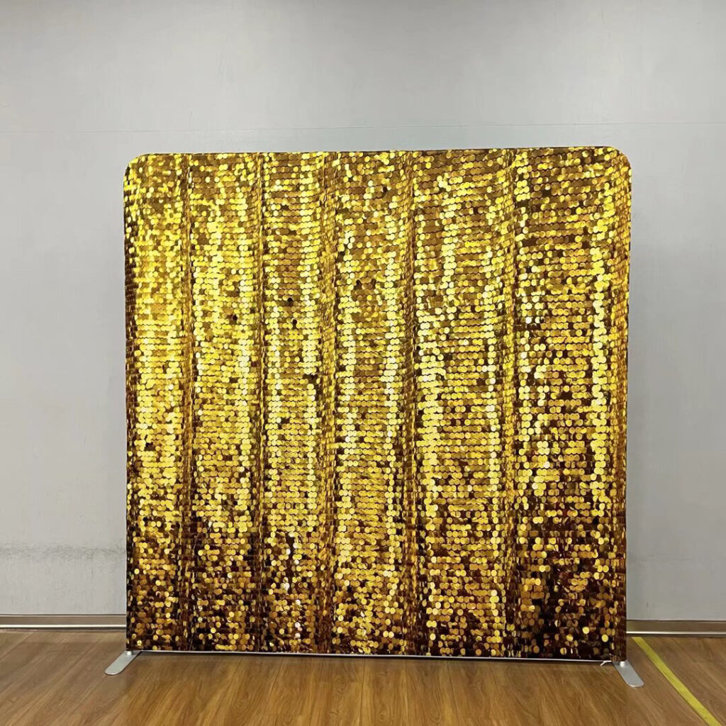 A shimmering gold sequin event backdrop standing on a wooden floor against a grey wall; contact us to book this glam photo background for your next party.