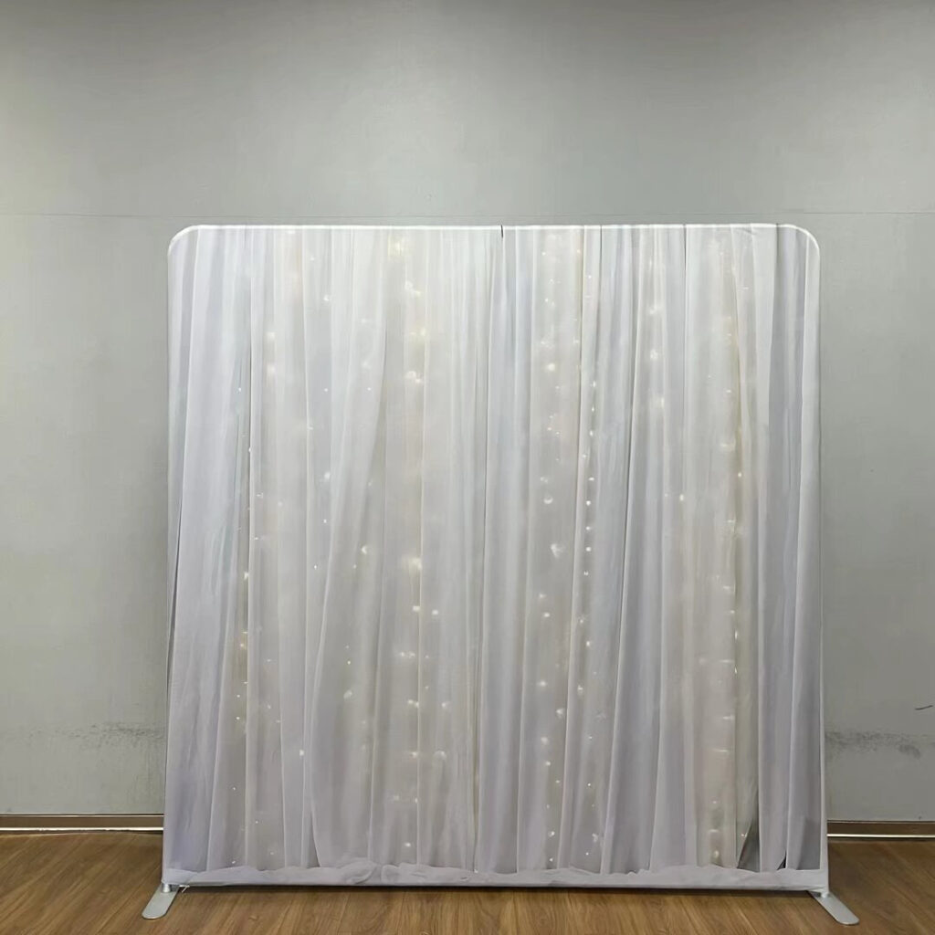 A large event backdrop featuring sheer white curtains draped in vertical folds with glowing warm fairy lights shining through from behind; contact us to book this ethereal and romantic background for your next party.