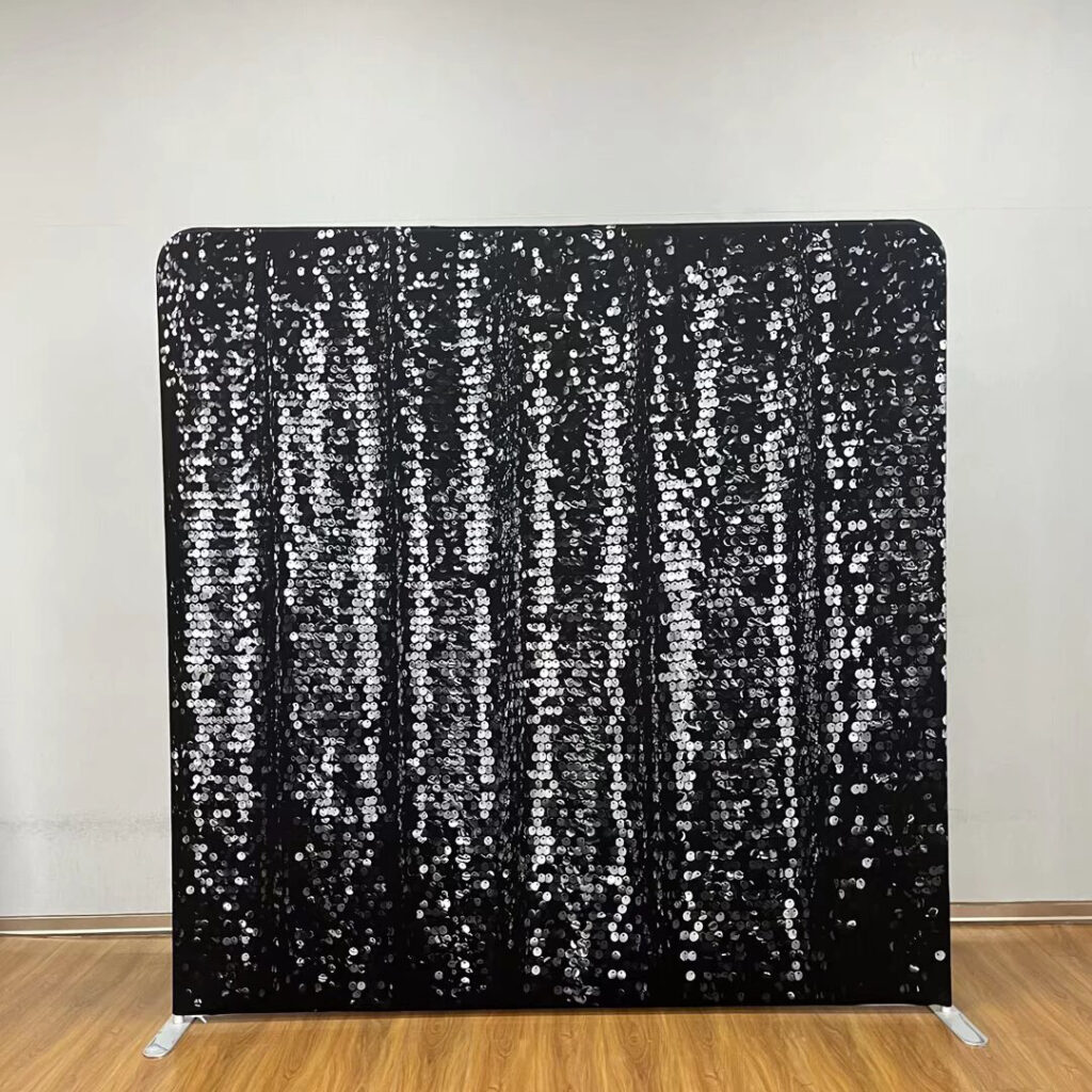 A shimmering black sequin event backdrop standing on a wooden floor; contact us to book this glamorous photo background for your next party.